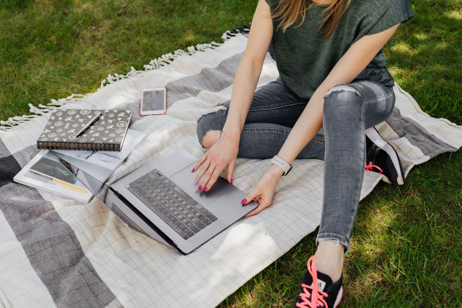From above of crop anonymous woman in casual clothes searching for information on laptop while sitting on blanket in sunny green park